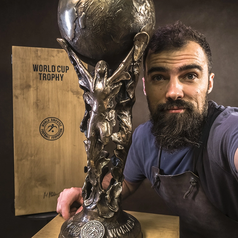 Puchar Mistrzostw Świata w Ampfutbolu Kobiet, wykonany z brązu, przedstawiający trzy piłkarki trzymające kulę ziemską.